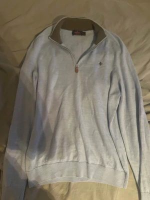 Ljusblå halvzip tröja från Morris  - Ljusblå långärmad tröja med halv dragkedja och ståkrage i kontrasterande beige. Tröjan har ribbade muddar vid ärmslut och nederkant samt en liten broderad logga på bröstet. Perfekt för lager på lager och enkel att matcha. Tröjan är storlek XL men passar både M och L jag är 178 väger 73 kg och den passar mig bra 