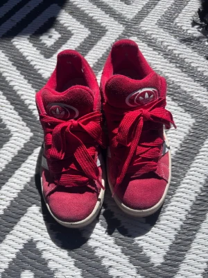 Röda Campus  - Snygga röda sneakers från Adidas med högt skaft och klassiska treklöver-loggan både fram och bak. Skorna har breda röda skosnören och vit sula. Ovandelen är i mockaliknande material och designen är sportig och retroinspirerad.