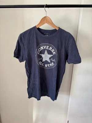 Mörkblå Converse All Star t-shirt - Snygg mörkblå t-shirt från Converse med det klassiska All Star-trycket i vitt framtill. T-shirten har rund hals och korta ärmar. Perfekt för dig som gillar streetwear och vill ha en enkel men ikonisk look.