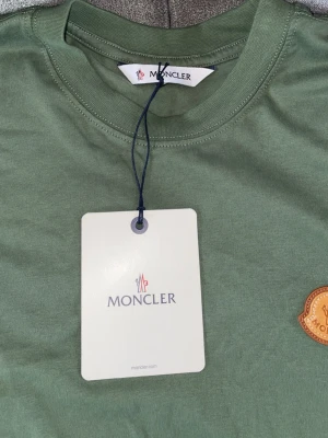 Grön Moncler t-shirt i bomull - Snygg grön t-shirt från Moncler med klassisk rund halsringning och korta ärmar. T-shirten har Monclers ikoniska logotyp i gult på bröstet och är tillverkad i mjuk bomull för en skön känsla. Perfekt till jeans eller shorts.