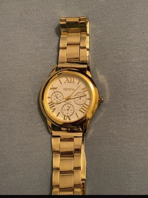 Guldfärgad klocka från Geneva - Stilren guldfärgad klocka från Geneva med metallarmband och vit urtavla. Klockan har romerska siffror, tre dekorativa subdials och klassisk design. Perfekt accessoar för dig som vill sticka ut med en elegant look.