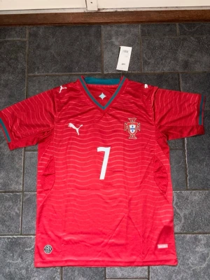 Portugal Ronaldo fotbollströja röd size S - Röd Portugal fotbollströja med nummer 7 och namnet Ronaldo på ryggen. Tröjan har gröna detaljer vid krage och ärmslut, vita Puma-loggor och Portugals landslagsemblem på bröstet. Tillverkad i lätt och ventilerande material, perfekt för fotbollsträning eller match. Med storlek d