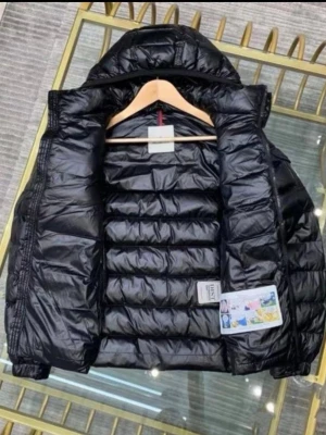 Svart dunjacka från Moncler - Snygg svart dunjacka från Moncler med glansig finish och horisontella sömmar. Jackan har huva och dragkedja framtill. Perfekt för kalla vinterdagar och ger en trendig look. Klassisk Moncler-logga på ärmen.