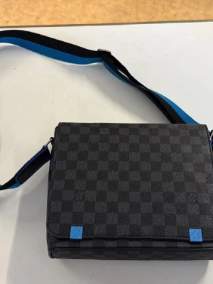 Svart axelväska med rutmönster - Snygg svart axelväska med diskret rutmönster och blå detaljer. Väskan har en justerbar axelrem i svart och blått, samt en praktisk lockstängning. Perfekt för dig som vill ha en stilren och modern look. Materialet är troligtvis konstläder eller canvas.