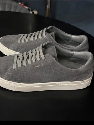Gråa sneakers från Axel Arigato - Stilrena sneakers i grå mocka från Axel Arigato med diskret logga på sidan. Skorna har vita sulor och matchande grå skosnören. Låg modell som passar perfekt till jeans 