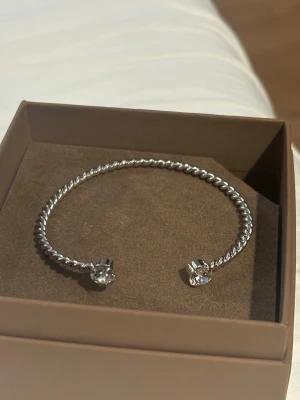 Silvrigt tvinnat armband med stenar från Caroline Svedbom - Snyggt armband i silverfärgad metall med tvinnad design och två gnistrande stenar i ändarna. Öppet och justerbart för enkel påtagning. Som Ny! Inga defekter! Pris kan diskuteras 