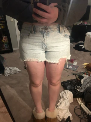 Ljusblå denimshorts med fransig kant - Säljer ett par ljusblå denimshorts med fransig, rå kant nertill. Shortsen har klassisk femficksmodell, knappgylf och är högmidjade. Perfekta för varma dagar och ger en avslappnad look.