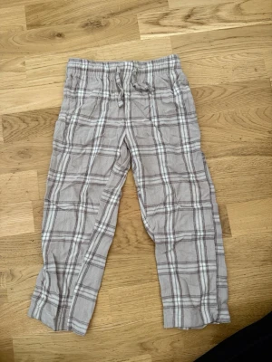 Rutiga pyjamasbyxor från 157 - Mysiga pyjamasbyxor i beige med rutigt mönster i grått och vitt från 157. Byxorna har resår och dragsko i midjan för extra komfort. Perfekta för chillkvällar hemma eller när du bara vill ta det lugnt.