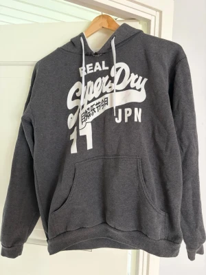 Mörkgrå Superdry hoodie - Säljer en mörkgrå hoodie från Superdry med stor vit logga och japansk text på bröstet. Hoodien har huva med vita snören, magficka och ribbade muddar vid ärmar och nederkant. Perfekt för en avslappnad och sportig stil. Har en liten defekt vid kragen.