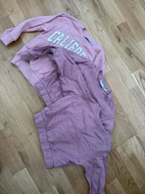 Två rosa sweatshirts, en med tryck - Säljer två rosa sweatshirts i barnstorlek. Ena tröjan har ett vitt 'CALIFORNIA'-tryck på bröstet och ribbade muddar, den andra är enfärgad och har en enkel, avslappnad passform. Båda har rund hals och är tillverkade i mjukt bomullsmaterial.