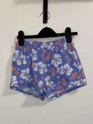 Blommiga blå shorts med resårmidja - Snygga blå shorts med vita, rosa och orange blommor. De har en bekväm resår i midjan och är perfekta för varma dagar. Lätta och luftiga med avslappnad passform och tropisk vibe.