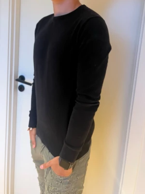 Svart basic långärmad tröja - En stilren svart långärmad tröja med rund halsringning. Tröjan har en enkel och klassisk design utan tryck eller mönster, perfekt för en clean look. Materialet ser mjukt och bekvämt ut, och passformen är rak med ribbade muddar vid ärmslut och nederkant. Modellen är 179
