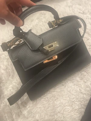 Svart handväska med gulddetaljer - Stilren svart handväska i strukturerat skinn med guldfärgade metalldetaljer och vridlås. Väskan har både handtag och avtagbar axelrem, perfekt för att bära på olika sätt. Klassisk och elegant design med lock och dekorativ rem över framsidan.