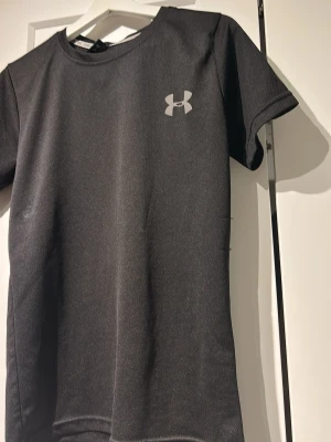 Svart tränings-tshirt från Under Armour - Snygg svart tränings-tshirt från Under Armour med klassisk logga på bröstet. Tillverkad i ett lätt och snabbtorkande funktionsmaterial som passar perfekt till gymmet eller löprundan. T-shirten har rund hals och korta ärmar.