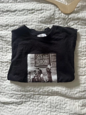 Svart t-shirt från bvaldi - Cool svart t-shirt med ett stort retroinspirerat tryck på bröstet där det står 'Birra Peroni' och visar en svartvit gatubild. Klassisk rund hals och korta ärmar. 