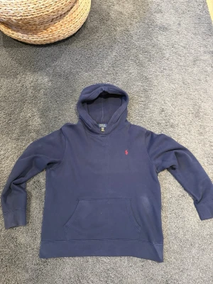 Mörkblå hoodie från Polo Ralph Lauren - Snygg mörkblå hoodie från Polo Ralph Lauren med klassisk huva och känguruficka. Ikoniska röda loggan broderad på bröstet. Tillverkad i mjuk bomull för skön känsla. Perfekt för en avslappnad och stilren look. Storlek XS skulle jag säga är själv 175 och väger runt 65kg och den är för liten på mig. Små ”fläckigt” som man ser på bilderna men det är endast sol blekt och inga mat fläckar. Syns inte lika tydligt irl.