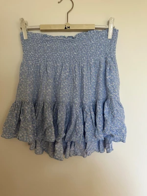 Blå volangkjol med blommönster - Söt blå kortkjol med små vita blommor och volangkant. Kjolen har smock i midjan för skön passform och är perfekt för varma dagar. Lätt och luftig.