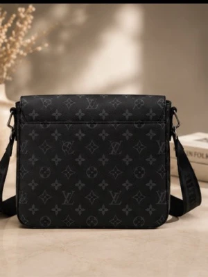 Väska  - Stilren svart axelväska i fyrkantig form med justerbar rem och klassiskt monogrammönster över hela väskan. Väskan är tillverkad i ett slätt material som liknar läder och har diskreta detaljer för en modern look.