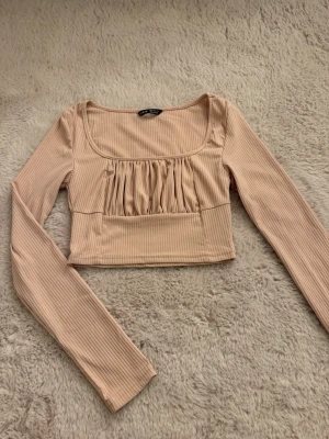 Långärmad topp - Trendig ljusrosa (ser beige ut i bilden men den är ljusrosa) croppad topp med långa ärmar och ribbad struktur. Toppen har en fyrkantig ringning och snygga rynkade detaljer framtill som ger en unik look. 