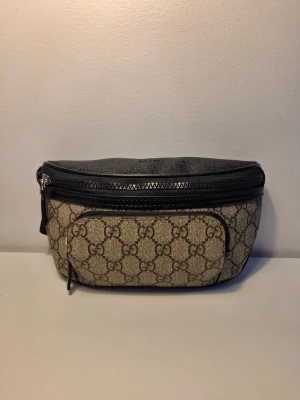 GG Beige axelväska  - Snygg beige axelväska från GG med klassiskt GG-monogram i brunt och svarta detaljer. Väskan har en framficka med dragkedja och justerbar rem. Tillverkad i canvas med läderdetaljer. Perfekt för dig som gillar streetstyle och vill ha något trendigt. Pris går att diskuteras! Levereras samma dag som köpet!