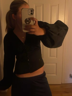 Svart blus med puffärm från Gina Tricot - Snygg svart blus från Gina Tricot i storlek XS från 14+! Blusen har markerad midja, v-ringning och stora puffiga långärmar som ger en trendig siluett. Knäppning framtill och mjukt, tunt tyg som känns skönt mot huden.