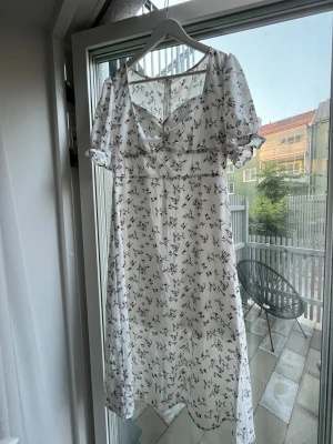 Vit blommig långklänning med puffärm - Söt vit långklänning med små blommor i rosa och grönt. Klänningen har puffärmar, v-ringning och lätt volang nedtill. Perfekt för sommarens alla tillfällen och ger en romantisk vibe.
