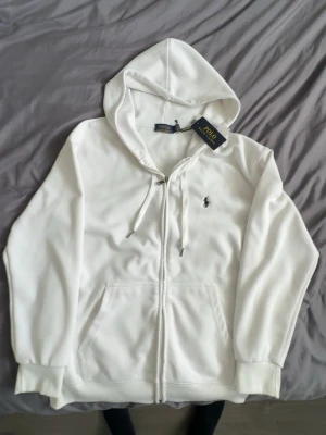 Vit hoodie från Polo Ralph Lauren - Snygg vit hoodie från Polo Ralph Lauren med dragkedja, huva med dragsko och klassisk broderad logga på bröstet. Tillverkad i mjukt material och har känguruficka framtill. Perfekt för en clean och stilren look. PASSAR S OCKSÅ.