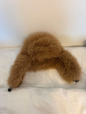 Brun fluffig pälsmössa med öronlappar - Säljer en supermjuk och fluffig brun pälsmössa med långa öronlappar. Mössan har en klassisk trappermodell och är perfekt för kalla dagar. Materialet är fuskpäls som ger en lyxig och varm känsla. Inga synliga märken på mössan.