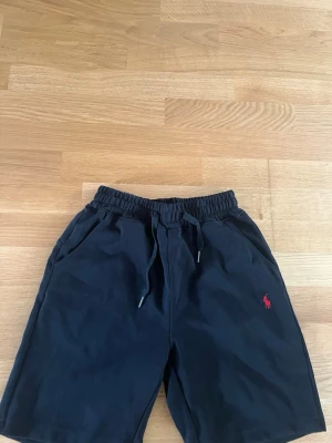 Svarta shorts från Polo Ralph Lauren - Svarta shorts från Polo Ralph Lauren med elastisk midja och snörning. Klassisk röd logga broderad på vänster ben. Tillverkade i mjuk bomull, perfekta för sommardagar. Två sidofickor och avslappnad passform.