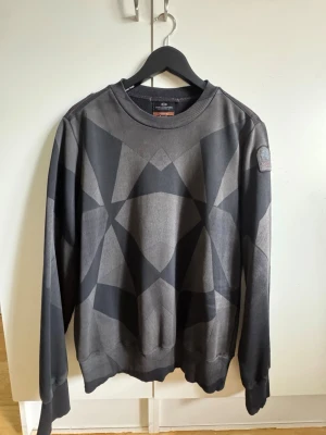 Parajumpers K2 Geomat (M) (Äkta) - Mörkgrå sweatshirt från Parajumpers med geometriskt mönster i olika nyanser av grått. Tröjan har rund halsringning, ribbade muddar och en patch med Parajumpers-logga på ärmen. Perfekt för en avslappnad streetwear-look.