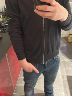 Svart ziptröja från Stone Island - Säljer en svart ziptröja från Stone Island med ikonisk patch på vänster ärm. Tröjan har ribbade muddar, hög krage och dragkedja framtill. Perfekt till jeans och enkel att styla för en clean look. Pris kan minska vid snabb affär.