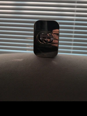 Gucci Guilty parfym metallisk flaska - Gucci Guilty parfym i en elegant, metallisk och rektangulär flaska med den ikoniska GG-loggan på framsidan. Flaskan har en mörk, spegelblank finish och insidan visar en djup röd vätska. Perfekt för dig som gillar lyxiga och stilrena dofter.