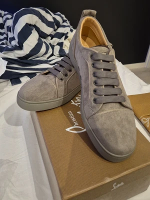 Grå mockasneakers från Christian Louboutin - Snygga grå sneakers från Christian Louboutin i mocka med klassisk röd sula och diskret logga. Perfekta för dig som vill ha en clean och lyxig look.