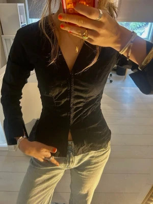 Svart figursydd blus från Weekday - Snygg svart blus från Weekday med figurnära passform och v-ringning. Blusen har långa ärmar med rynkade detaljer längs sidorna och ärmarna. Tillverkad i ett tunt, lätt material som ger en cool och modern look.