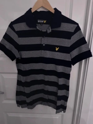 Randig pikétröja från Lyle & Scott - Snygg pikétröja från Lyle & Scott med breda marinblå och grå ränder. Klassisk krage, korta ärmar och knappslå framtill. Tillverkad i mjuk bomull och har den ikoniska gula loggan broderad på bröstet.
