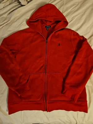 Röd hoodie från Polo Ralph Lauren - Röd hoodie från Polo Ralph Lauren med dragkedja framtill och klassisk huva. Hoodien har två fickor och den ikoniska lilla loggan broderad på bröstet. Perfekt för dig som gillar en enkel men snygg stil.