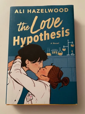The Love Hypothesis Collector’s Edition  - Säljer romanen 'The Love Hypothesis' av Ali Hazelwood. Boken har ett blått och gult omslag med tecknad illustration av två personer som kysser varandra i ett labb. Sidorna har rosa kanter med vita vetenskapliga symboler. Inbunden och på engelska. Helt ny, aldrig läst