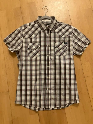 Rutig kortärmad skjorta från Emilio herr storlek L - Kortärmad skjorta från Emilio i vitt, blått och rött rutigt mönster. Skjortan har två bröstfickor med lock och knappar samt klassisk krage. Tillverkad i bomull för en skön känsla. Perfekt för en avslappnad och stilren look. Snygg till sommaren. Jätte fin skick, herr storlek L