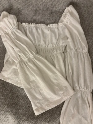 Vit blus med volang - Säljer en söt vit blus som kan bäras normalt eller som offshoulder med volang och långa puffärmar. Blusen har resår vid axlar och midja för en snygg passform och är tillverkad i ett lätt, luftigt tyg som känns perfekt för varmare dagar.