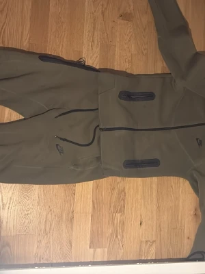 Olivgröna mjukisbyxor från Nike - Säljer ett par olivgröna mjukisbyxor från Nike med svarta dragkedjor och fickor. Byxorna har en sportig look med resår i midjan och dragsko för bästa passform. Perfekta för chill eller träning, tillverkade i mjukt och stretchigt material. Nike tech som har ett litet problem vid foten som är sytt.