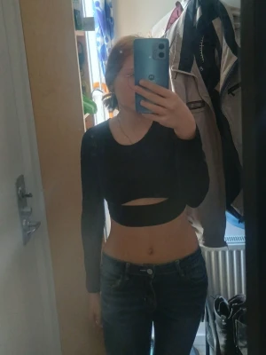 Svart croppad topp med snörning - Trendig svart croppad topp med lång ärm och cool cut-out framtill. Baksidan har öppen rygg med snygg snörningsdetalj som ger en edgy vibe. Perfekt för dig som vill sticka ut och matcha med höga jeans eller kjol.