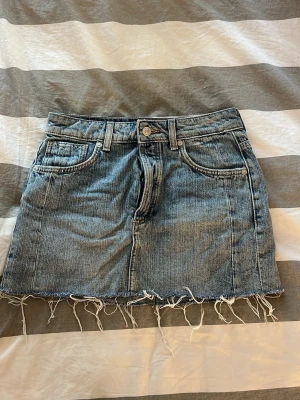 Blå jeanskjol med fransig kant - Säljer en blå jeanskjol i klassisk modell med fem fickor och rå, fransig kant nedtill. Kjolen har bälteshällor och knappstängning framtill. Perfekt för dig som gillar en avslappnad och trendig stil.