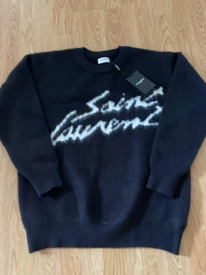 Svart Saint Laurent stickad tröja - Snygg svart stickad tröja från Saint Laurent med vit logga i borstigt garn framtill. Tröjan har rund halsringning, långa ärmar och ribbade muddar. Perfekt för dig som vill ha en trendig och exklusiv look.