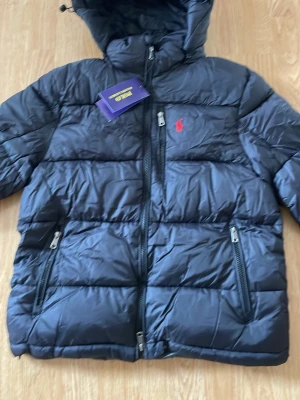 Polo Ralph Lauren jacka - Snygg svart pufferjacka från Polo Ralph Lauren med huva och två dragkedjeförsedda fickor framtill. Jackan har det klassiska röda Polo-logotypen broderad på bröstet och quiltad design. Perfekt för kalla vinterdagar.