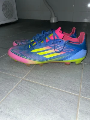 Adidas f50 celestial victory - Säljer ett par riktigt snygga fotbollsskor från Adidas i blått, rosa och gult. Skorna har klassiska tre ränder på sidan, snörning och en låg profil. Ovandelen är i syntetmaterial och sulan har fasta dobbar, perfekta för spel på gräsplan.