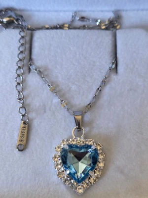 Halsband med blå hjärtformad sten - Snyggt halsband i silverfärgat stål med en stor, blå hjärtformad sten som hänge. Runt stenen sitter gnistrande klara pärlor som ger extra bling. Kedjan har ett elegant mönster och justerbar längd. Perfekt för dig som vill sticka ut med ett statement-smycke.