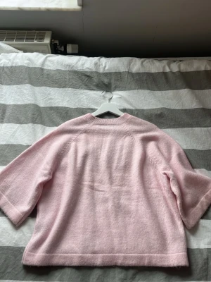 Ljusrosa stickad tröja med vida ärmar - Mysig ljusrosa stickad tröja med trekvartsvida ärmar och rund halsringning. Tröjan har en avslappnad passform och är tillverkad i ett fluffigt, mjukt material som känns skönt mot huden. Perfekt för lager-på-lager och en söt look.