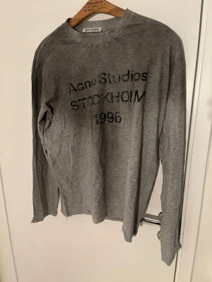 Grå långärmad tröja Acne Studios - Säljer en grå långärmad tröja från Acne Studios med svart text 'Acne Studios STOCKHOLM 1996' framtill. Tröjan har en enkel, rak passform och är tillverkad i mjuk bomull. Perfekt för en avslappnad och stilren look.