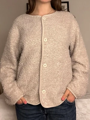 Beige teddyjacka med knappar🤍 - Mysig beige cardiganjacka i teddy-material med rund halsringning och fyra vita knappar framtill. Jackan har avslappnad passform och långa ärmar. Perfekt för dig som gillar en enkel men trendig look.🤍