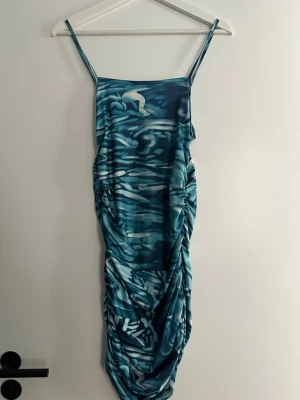 Blå mönstrad klänning från SHEIN - Trendig blå klänning från SHEIN med vattenliknande mönster och tunna axelband. Klänningen har åtsittande passform och rynkade sidor som ger en snygg siluett. Tillverkad i ett mjukt, stretchigt material som känns bekvämt mot huden.